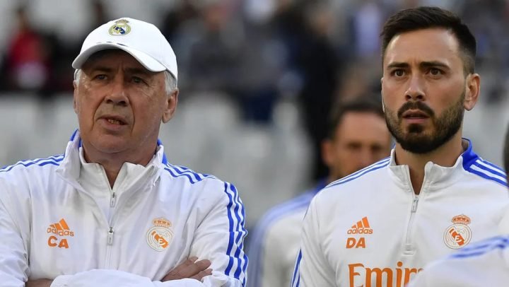Ảnh con trai Davide Ancelotti ngồi ghế cạnh cha mình, HLV Carlo Ancelotti, tại một buổi họp báo hoặc trong khu vực kỹ thuật, thể hiện sự học hỏi và chuyển giao thế hệ trong công việc.